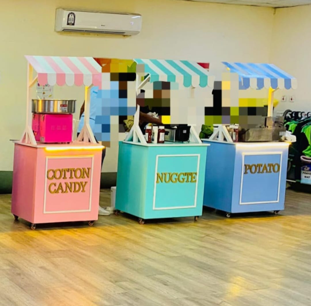 Three colorful mobile food and candy carts on a wooden floor in an indoor room. The pink cart is labeled 'COTTON CANDY' with a pink-and-white striped awning and a cotton candy machine.